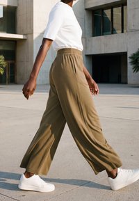 Personne marchant à l'extérieur près d'un bâtiment moderne en béton, portant des baskets blanches, un pantalon olive à jambes larges et un haut blanc à manches courtes.