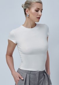 Camiseta blanca ajustada de manga corta y escote redondo. Presenta un sutil logo bordado en el pecho. Tejido de algodón suave.