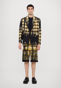Black and yellow plaid blazer with a splash pattern, paired with matching shorts. Model wears black shoes and a simple black shirt underneath.