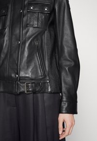 Černá kožená bunda s patentkami, dvěma předními kapsami, vertikálním žebrováním po stranách a páskem v oblasti pasu pro nastavitelnou velikost.