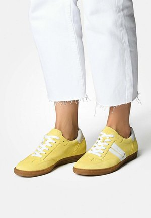 Yellow suede sneakers feature a white stripe, white laces, and a rubber sole. Paired with frayed white jeans for a casual look.