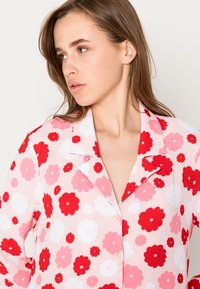 Camisa de pijama floral com fundo rosa, flores vermelhas e brancas, gola com pico e fecho de botão. Tecido texturado.