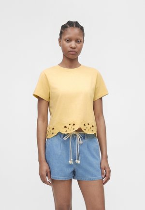 Femme aux cheveux tressés portant un haut jaune à manches courtes avec des découpes florales et un short en jean bleu avec une taille à cordon.