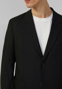 Schwarzer Blazer mit Reverskragen, Ein-Knopf-Verschluss und zwei Taschen. Über einem weißen Rundhals-T-Shirt getragen, glatte Stofftextur.