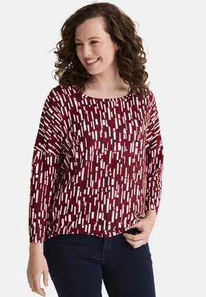Maroon long-sleeve top featuring a geometric white pattern, round neckline, and relaxed fit, paired with dark blue jeans.