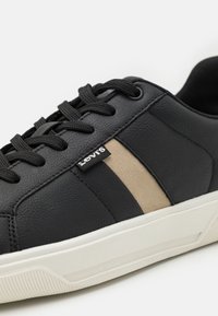 Zapatilla de cuero negra con franjas beiges, superficie texturizada, suela blanca en contraste, cordones negros y un pequeño logo en el lateral.