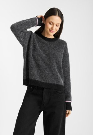 Grijze, fuzzy gebreide trui met een zwarte kraag, voorzien van roze accenten bij de manchetten en een relaxed fit. Gepaard met zwarte broeken.