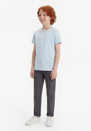 Lächelnder rothaariger Junge, der ein hellblaues T-Shirt, dunkelgraue Hosen mit aufgekrempelten Beinenden und weiße Turnschuhe trägt, steht vor einem weißen Hintergrund.