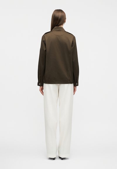 Γυναίκα με μακριά ίσια μαλλιά που φοράει σκούρο καφέ σακάκι και λευκό παντελόνι φαρδιάς γραμμής, στέκεται με την πλάτη στραμμένη σε απλό φόντο.