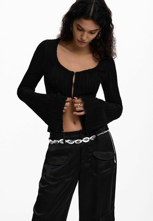 Donna con capelli scuri e mossi che indossa un top corto nero a maniche lunghe con texture e pantaloni neri con una cintura a catena argento, mentre sistema il suo top.