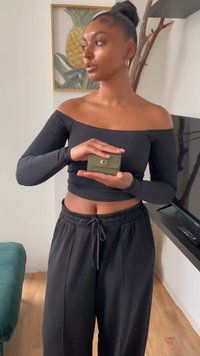 Small olive green handbag with a gold clasp, held in hands. The setting features a black long-sleeve top and loose black pants.