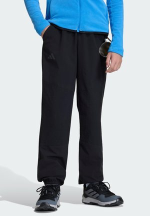 Person trägt schwarze Adidas-Hose, schwarze und graue Adidas-Sneaker sowie eine blaue Jacke und hält einen Metallbecher in der Hand.