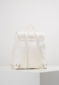 Vit faux läder ryggsäck med dubbla remmar och ett handtag upptill. Har en slät yta och minimalistisk design. Inga synliga detaljer.