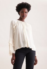 Blusa bianca plissettata con maniche a volant, scollatura rotonda e vestibilità rilassata, abbinata a jeans scuri per creare contrasto. Texture del tessuto morbido visibile.