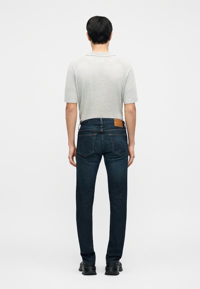 Γκρι κοντομάνικο πουκάμισο, σκούρο μπλε τζιν τζιν με δερμάτινη ετικέτα στη μέση, και μαύρα παπούτσια. Θέα από πίσω.