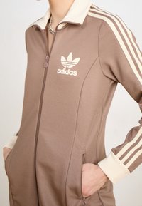 Brun zip-up träningsoverall med krämfärgade detaljer, med det distinkta Adidas-logotypen och tre vita ränder längs ärmarna. Slät textur.