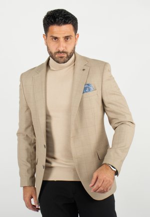 Blazer à carreaux beige en tissu texturé, associé à un col roulé beige clair et un pochette de costume bleue à motifs. Un pantalon noir complète la tenue.