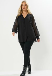 Camicia nera con bottoni, maniche lunghe trasparenti, colletto arricciato e dettagli decorativi con bottoni sui polsini. Abbinata a pantaloni neri aderenti e stivaletti.