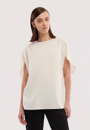 Junge Frau mit glatten braunen Haaren, die ein locker sitzendes weißes kurzärmeliges Oberteil und schwarze Hosen trägt, vor einem schlichten Hintergrund stehend.