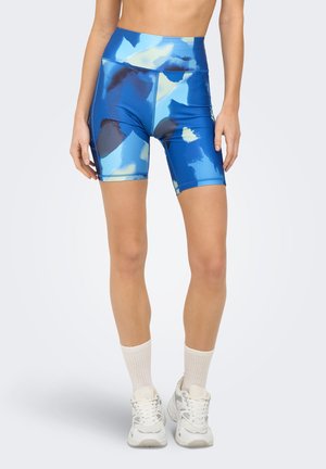 Femme porte un short de vélo bleu taille haute à imprimé abstrait, des chaussettes blanches montantes et des baskets blanches épaisses, debout devant un fond uni.