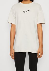 Krämfärgad bomulls t-shirt med svart Nike swoosh-logotyp på bröstet. Har korta ärmar och en avslappnad passform.