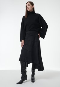 Zwarte oversized coltrui gecombineerd met een gelaagde zwarte rok met horizontale strepen. Afgemaakt met hoge zwarte laarzen tot de knie.