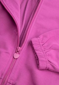 Różowa kurtka zapinana na zamek z teksturowanym wnętrzem, z suwakiem w kształcie serca i marszczonymi mankietami na rękawach.