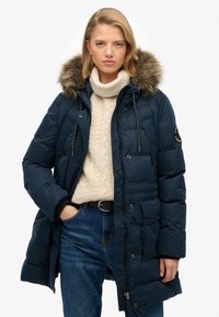 Giacca a vento blu navy imbottita con cappuccio in pelliccia sintetica, chiusura con zip e bottoni a pressione, texture trapuntata, numerose tasche e dettagli con cordino. Indossata sopra un maglione crema.