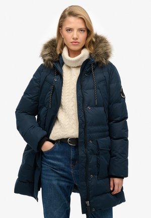 Marineblå pufferjakke med faux pels hætte, lynlås og trykknaplukning, quiltet tekstur, flere lommer og snoretræk. Båret over en lys cremefarvet sweater.