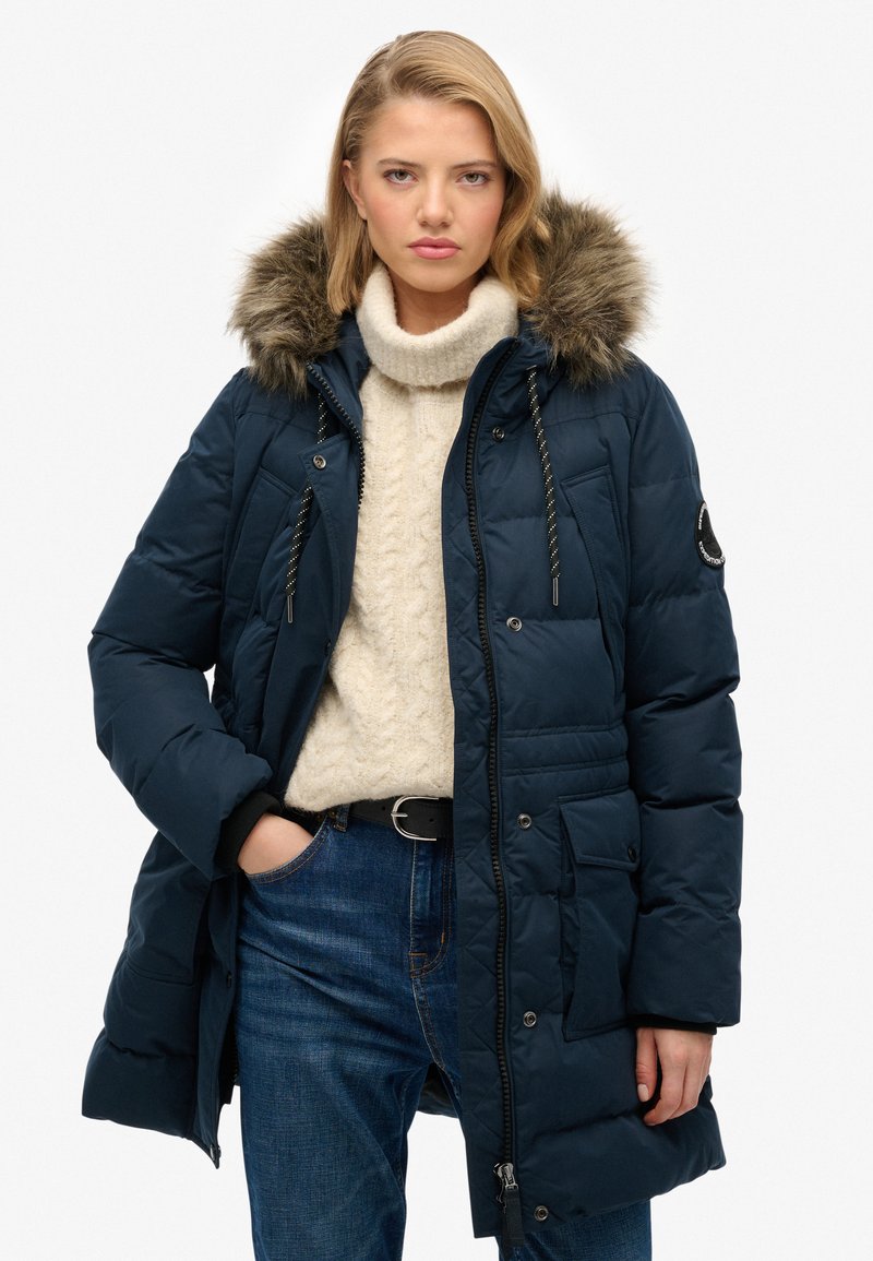 Giacca a vento blu navy imbottita con cappuccio in pelliccia sintetica, chiusura con zip e bottoni a pressione, texture trapuntata, numerose tasche e dettagli con cordino. Indossata sopra un maglione crema.