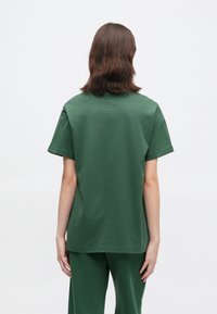 Άτομο με καστανά μαλλιά μέχρι τους ώμους στέκεται με πλάτη, φορώντας ένα απλό πράσινο κοντομάνικο μπλουζάκι και αντίστοιχες πράσινες παντελόνες.