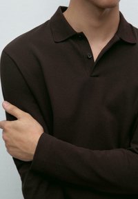 Man in dark brown long-sleeve polo shirt crossing his left arm over his chest, head partially visible against plain background.