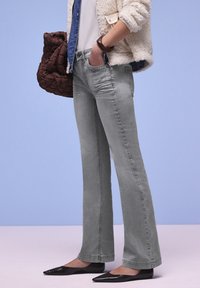 Personne portant un jean évasé gris clair, des chaussures plates noires pointues, des hauts superposés, tenant un sac matelassé marron sur fond bleu.