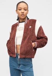 Νεαρός με κοτσίδες, φορώντας ένα μοβ και άσπρο χνουδωτό μπουφάν, εκρού κοντό τοπ με λογότυπο ήλιου και μπλε τζιν, με τα χέρια στις τσέπες.