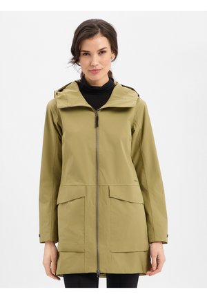 Frau trägt eine lange olivgrüne Kapuzenjacke mit vorderem Reißverschluss und großen Pattentaschen, steht vor einem weißen Hintergrund.