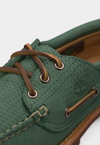 Πράσινο δερμάτινο παπούτσι τύπου boat με καφέ δερμάτινα κορδόνια περασμένα από μεταλλικά αγκράφια και μπεζ ραφές σε καφέ σόλα.