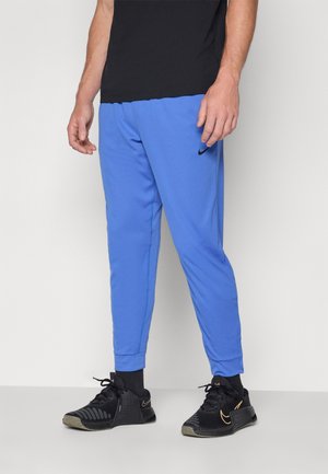 Uomo che indossa pantaloni da jogging Nike blu brillante, calzini neri, sneakers Nike nere e oro, e una maglietta nera, in piedi su un pavimento grigio.