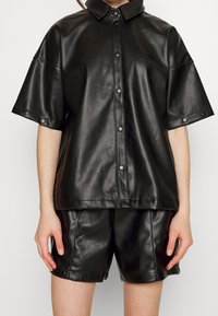 Svart faux läderskjorta med korta ärmar, tryckknappslås framtill och matchande shorts. Slät textur med en subtil lyster.