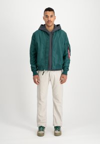 Groene bomberjack met een rits en geribde manchetten, gedragen over een grijze hoodie. Lichtgekleurde broek en groene sneakers maken de outfit compleet.