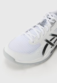 Chaussure de sport blanche avec une tige en mesh, une semelle en caoutchouc et des accents noirs. Le design comprend une languette rembourrée et des lacets pour un maintien sécurisé.