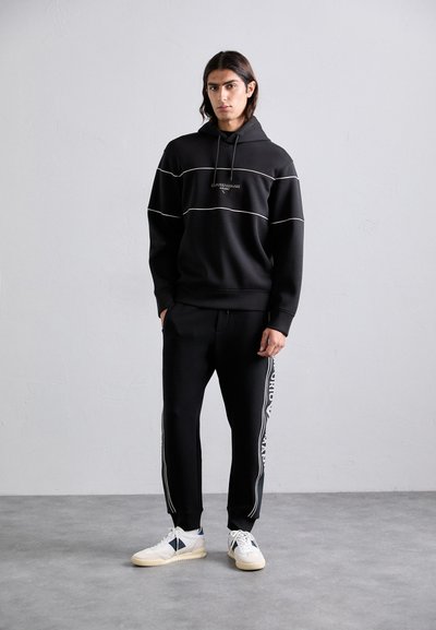 Homem em pé contra uma parede lisa, vestindo um hoodie preto com finas riscas brancas e calças de fato de treino pretas combinando com texto branco na lateral, tênis brancos.