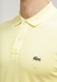 Polo majica v svetlo rumeni barvi z teksturirano tkanino, ki ima tri gumbke in zeleno vezeno krokodilovo logo na levem prsnem delu.