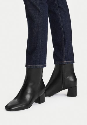 Czarne skórzane botki na kostkę z kwadratowym noskiem i masywnym obcasem. Wyposażone w boczny zamek błyskawiczny o gładkiej fakturze, zestawione z ciemnymi jeansami.