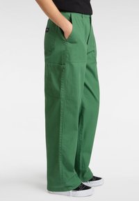 Personne portant un pantalon cargo vert ample avec des poches latérales et arrière, les mains dans les poches, associé à des baskets noires et blanches.