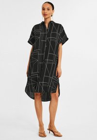 Schwarzes Hemdkleid mit kurzen Ärmeln, das ein geometrisches Muster aus weißen Linien aufweist, eine Knopfleiste hat und seitliche Schlitze für mehr Bewegungsfreiheit bietet.