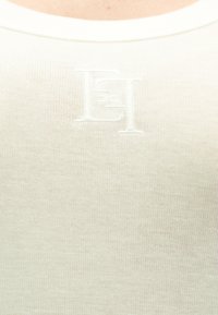 Λευκό πλεκτό αμάνικο τοπ με στρογγυλή λαιμόκοψη και διακριτική υφή υφάσματος, με ανάγλυφο λογότυπο στο κέντρο.