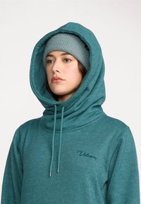 Jeune femme portant un sweat à capuche teal avec cordons, sous un bonnet en laine gris, regardant sur le côté contre un fond uni.