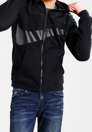 Osoba ubrana w czarną bluzę z zamkiem i dużym ciemnoszarym logo Swoosh oraz niebieskie dżinsy, poprawiająca bluzę przy szyi.