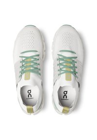 Baskets de course blanches avec tige en mesh, lacets verts et accents jaunes. Présente un logo sur la languette et des zones perforées pour la respirabilité.