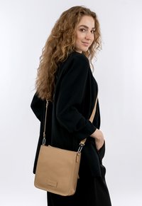 Borsa in pelle beige con finitura testurizzata, forma quadrata, tracolla regolabile e dettagli con logo, indossata sulla spalla.